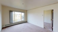 This single-floor space features a clean, minimalist design with neutral tones throughout. The main living area is finished with wall-to-wall carpet in a light beige hue, complementing the cream-colored walls and white ceiling, creating a warm and cohesive aesthetic. A large sliding window dominates one wall, flooding the room with natural light and providing views of the surrounding neighborhood, including mature trees and adjacent buildings. The window is equipped with a baseboard heating unit beneath it, offering an efficient and unobtrusive climate control solution. The open wall space provides ample flexibility for furniture arrangement, while a doorway leads to an adjoining room, suggesting a functional layout designed for ease of movement. The ceiling is smooth and flat, contributing to the room's clean, understated character. The overall ambiance is calm and neutral, offering a versatile blank canvas well-suited for a variety of uses. The space benefits from good natural light, an elevated position with outward views, and practical heating infrastructure, making it a comfortable and functional environment.