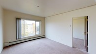 This single-floor space features a clean, minimalist design with neutral tones throughout. The main living area is finished with wall-to-wall carpet in a light beige hue, complementing the cream-colored walls and white ceiling, creating a warm and cohesive aesthetic. A large sliding window dominates one wall, flooding the room with natural light and providing views of the surrounding neighborhood, including mature trees and adjacent buildings. The window is equipped with a baseboard heating unit beneath it, offering an efficient and unobtrusive climate control solution. The open wall space provides ample flexibility for furniture arrangement, while a doorway leads to an adjoining room, suggesting a functional layout designed for ease of movement. The ceiling is smooth and flat, contributing to the room's clean, understated character. The overall ambiance is calm and neutral, offering a versatile blank canvas well-suited for a variety of uses. The space benefits from good natural light, an elevated position with outward views, and practical heating infrastructure, making it a comfortable and functional environment.