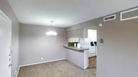 This single-floor space showcases an open-concept design that seamlessly connects the dining area and kitchen. The dining area is finished with plush carpeting and neutral grey-taupe walls, and is illuminated by a classic multi-bulb chandelier that serves as a focal point overhead. A textured ceiling adds subtle depth to the space.

The kitchen is defined by a peninsula with a laminate countertop, providing both workspace and a casual bar-style seating option. White cabinetry lines the upper portion of the kitchen, complemented by white appliances including a refrigerator and range. The kitchen transitions to a hard flooring surface, offering a practical contrast to the carpeted dining area. A pass-through opening connects the kitchen to an additional area beyond, enhancing the sense of flow throughout the unit.

The overall ambiance is clean and understated, with a consistent neutral color palette tying the spaces together. Overhead ventilation and a visible smoke detector reflect functional, well-considered details throughout the layout.