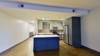 This single-floor unit offers a thoughtfully arranged layout that maximizes its generous footprint. The open-plan kitchen and living area serves as the heart of the space, featuring rich dark hardwood-style flooring that extends throughout. The kitchen is outfitted with gray shaker-style cabinetry, white quartz countertops, a textured tile backsplash, stainless steel appliances including a full-size refrigerator, and a striking cobalt blue center island that doubles as a bold design focal point. Glass open shelving adds a modern touch near the sink area. A long hallway connects the main living space to the private areas of the unit, lined with louvered doors and recessed lighting overhead. The bedroom offers a comfortable, quiet retreat with carpeted flooring and a dedicated walk-in closet equipped with built-in shelving and hanging rods on multiple walls, providing ample storage. The bathroom is a standout feature, fully clad in white subway tile with gray accent inserts, and includes a deep soaking tub, a separate shower area with a glass enclosure, and clean, polished fixtures throughout. The overall aesthetic is contemporary and cohesive, with a consistent cool-toned gray and blue palette.