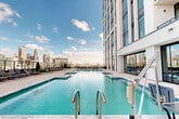 Located at 615 E Morehead St in Charlotte, NC, this rooftop amenity level offers a sophisticated outdoor retreat elevated above the city. At its centerpiece is a generously sized resort-style swimming pool with crystal-clear water, flanked by a surrounding deck finished in large-format stone pavers and accented with manicured planters and lounge seating. The pool deck provides direct, unobstructed views of the Charlotte skyline, creating a striking urban backdrop.

The outdoor space extends well beyond the pool, encompassing multiple distinct lounging areas thoughtfully designed for both relaxation and socializing. Covered pergola-style cabanas feature decorative lattice screens, pendant lighting, and plush upholstered seating arranged for intimate gatherings. Open-air seating areas along the glass-railed perimeter offer panoramic views of the surrounding cityscape, furnished with woven lounge chairs and accent tables. Additional zones include an outdoor dining area with a built-in grilling station, ideal for alfresco entertaining.

Wide, tiled walkways connect each section of the deck, running alongside floor-to-ceiling glass curtain walls of the adjacent tower. The overall design balances modern architectural elements with lush greenery, creating a polished and inviting outdoor environment in the heart of Charlotte.