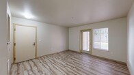 This single-floor residential unit offers a clean, functional layout finished consistently with light hardwood-style flooring throughout every room, lending a bright and cohesive feel to the entire space.

The living area is generously proportioned, featuring neutral painted walls with warm wood-toned door and window trim. A glass-panel exterior door opens to an outdoor view, while a large adjacent window allows ample natural light to fill the room, creating an open and airy atmosphere.

The kitchen is efficiently designed with light wood cabinetry, grey laminate countertops, a double-basin sink, and a full suite of white appliances including a refrigerator, stove, dishwasher, and microwave. A pass-through window connects the kitchen to the main living area, enhancing the sense of openness.

The bedroom is a well-sized room equipped with a sliding-door closet featuring built-in shelving, providing practical storage. It connects conveniently to an adjoining bathroom. A second bedroom or flex room is also present, similarly outfitted with a sliding closet door.

Two bathrooms serve the unit — one featuring a tub/shower combination with accessibility grab bars and a vanity with countertop, and a second full bathroom with a tub/shower combo and standard fixtures. Both bathrooms continue the light flooring found throughout the unit.