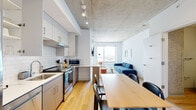 Located at 1456 N Dayton St in Chicago's vibrant River West neighborhood, this contemporary single-floor unit showcases a clean, modern aesthetic defined by exposed concrete ceilings, warm hardwood flooring, and an abundance of natural light. The open-plan living and dining area flows seamlessly from the kitchen, creating an inviting, functional space. Floor-to-ceiling windows frame sweeping city views, filling the living room with light and offering a striking urban backdrop. The living area features a wall-mounted flat-screen TV, modern furnishings, and colorful framed artwork that adds personality to the neutral palette. The well-appointed kitchen is equipped with stainless steel appliances, white subway tile backsplash, gray cabinetry, white countertops, and ample storage. A dedicated dining area with a large wood table provides comfortable seating for multiple guests. The bedroom is a tranquil retreat with hardwood floors, a built-in closet, and a window overlooking the cityscape, accented by an exposed concrete column. Two bathrooms feature gray vanities, white quartz countertops, subway-tiled walk-in showers, and sliding glass or rod-hung enclosures. The space balances industrial architectural elements with a refined, contemporary interior throughout.