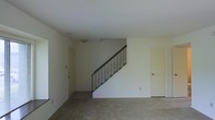 This two-story space presents a straightforward and functional layout centered around a carpeted main living area on the ground floor. The room is finished with neutral-toned walls and a smooth white ceiling, creating a bright and clean atmosphere. A staircase with a dark metal railing runs along the interior wall, providing access to the upper floor. The staircase serves as a subtle architectural focal point within the otherwise understated space. Two doors are visible on the main level — one along the left wall and another on the right side of the room — suggesting access to additional rooms or storage areas. The carpeting throughout the main floor adds a sense of warmth and comfort underfoot. Recessed ventilation details are visible in the ceiling, and a small light fixture is mounted near the base of the staircase landing. The overall ambiance is minimal and move-in ready, with a neutral palette that lends itself well to a variety of interior styles. The open feel of the main level, combined with the vertical access to a second floor, makes this a practical and adaptable multi-level space.