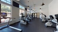 This fitness center offers a spacious, single-floor layout designed to accommodate a full range of cardio and exercise activities. The open floor plan is finished with durable dark rubber flooring throughout, providing a clean, functional surface well-suited for high-traffic gym use.

The main workout area is lined with a generous selection of TRUE brand elliptical trainers, treadmills, and stair climbers, arranged to maximize space and accessibility. A dedicated section houses multiple recumbent stationary bikes, catering to low-impact exercise needs. Additional equipment includes a StairMaster and a rack of exercise balls and accessories along the perimeter wall.

Natural light fills the space through large, multi-pane windows with bold black frames, offering views of an outdoor pool area and surrounding greenery — creating an inviting atmosphere for workouts. Clerestory windows near the roofline further enhance brightness and contribute to the room's elevated, airy feel.

Overhead, a series of industrial-style pendant lights with dark dome shades provide warm, focused illumination throughout the space. Wall-mounted flat-screen televisions are positioned at multiple vantage points for convenience. A textured ceiling adds subtle architectural character, while white walls keep the environment clean and energizing. Entry is provided through both a standard door and a set of double glass-panel doors.