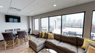 This contemporary rooftop amenity space seamlessly blends indoor comfort with outdoor urban living. The interior lounge area is thoughtfully designed with a large tufted sectional sofa accented by yellow throw pillows, complemented by a dining table and chairs — ideal for casual gatherings. A drop ceiling with recessed lighting and warm wood-tone flooring add a polished, modern finish throughout.

Floor-to-ceiling windows and a sliding glass door flood the interior with natural light while framing sweeping views of the surrounding city skyline. The transition to the outdoor terrace is effortless, opening onto a generous rooftop deck equipped with multiple gas grills, outdoor dining sets, and lounge seating arranged to take full advantage of the elevated cityscape. The railing-lined perimeter ensures unobstructed sightlines across the urban horizon.

The overall ambiance strikes a balance between a relaxed residential feel and a stylish, functional communal space. Whether used for entertaining, casual relaxation, or al fresco dining, this rooftop lounge offers a compelling combination of thoughtful interior design and impressive outdoor amenities with a striking city backdrop.