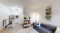 Located at 3030 W Sam Houston Pkwy S in Houston, TX, this single-floor unit offers a well-organized and thoughtfully designed layout that maximizes every inch of available space. The living area serves as the heart of the unit, featuring light hardwood-style flooring, a textured ceiling, and bright white walls that create an open, airy feel. A comfortable sofa, geometric area rug, and decorative touches — including gold hexagonal wall accents and a macramé plant shelf — lend the space a modern boho character. Natural light filters in through a window near the entry door, enhancing the welcoming ambiance. The kitchenette is efficiently designed with crisp white cabinetry, a mosaic tile backsplash, stainless steel refrigerator, and a compact sink. A sleek bar-height table with a wood top and black metal frame provides a practical dining or workspace. A contemporary multi-bulb chandelier overhead adds a stylish finishing touch. The bathroom is cleanly appointed with a wide marble-style countertop, under-mount sink, large mirror, and a shower-tub combination. White cabinetry and light flooring maintain the unit's cohesive, fresh aesthetic throughout. This space combines functional design with modern style in a convenient Houston location.