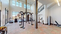 This two-story fitness center is designed to accommodate a wide range of workout needs within a spacious, well-lit environment. The main level immediately impresses with its dramatic double-height ceilings, supported by structural columns and flanked by large floor-to-ceiling windows and clerestory windows above, flooding the space with abundant natural light. Ceiling fans circulate air throughout the open volume. The ground floor houses a robust selection of equipment, including a multi-functional cable and suspension training rig, a heavy bag, cardio machines such as stair climbers and stationary bikes, and various strength training stations. A dedicated area near the entrance features built-in storage cubbies and an adjacent utility station. Framed athletic artwork lines the walls, contributing an energetic aesthetic. A staircase with a distinctive metal baluster railing leads to the second floor, which offers additional workout space. The upper level benefits from large windows overlooking the surrounding urban streetscape, providing an open and airy atmosphere. Additional cardio and strength equipment, including benches and cable machines, are positioned to take advantage of the views. Durable, multi-toned flooring runs throughout both levels, completing this well-equipped and architecturally dynamic fitness facility.