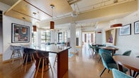 This thoughtfully designed single-floor communal space blends industrial and contemporary aesthetics to create a versatile, welcoming environment. Polished concrete floors flow throughout, complementing the exposed ceiling infrastructure complete with visible ductwork, pipes, and track lighting that lend the space an urban, loft-inspired character.

The open-plan layout encompasses several distinct seating zones. A central lounge area features plush sofas and leather armchairs arranged around a coffee table, anchored by a vibrant teal-toned area rug and framed by large, steel-framed windows that flood the room with natural light and offer views of surrounding greenery. Adjacent to this, multiple intimate dining clusters showcase teal velvet chairs paired with warm wood round tables, decorated with framed black-and-white artwork and decorative vases on built-in display shelves.

A bar-height island with dark wood accents and modern transparent stools serves as a casual gathering point, while a separate high-top bar area with black pedestal stools sits beneath a striking open staircase with a steel and glass railing, flanked by large abstract teal artwork. The space is further enhanced by warm pendant lighting in rich orange tones throughout.

A compact, well-appointed kitchenette with dark cabinetry, glass-front upper cabinets, a vibrant tiled backsplash, and stainless steel appliances provides convenient service support for the lounge areas.