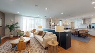 This thoughtfully designed single-floor space offers an open, flowing layout that seamlessly connects a fully equipped kitchen, a dining area, and a generously sized lounge. The kitchen is a standout feature, showcasing striking veined quartz countertops, navy cabinetry, a large center island with an undermount sink, stainless steel appliances including a built-in double oven and refrigerator, and a sleek stainless steel range hood set against a dramatic marble-look backsplash. Warm hardwood flooring runs throughout, tying the spaces together with a cohesive, inviting feel. The lounge area is anchored by a plush cream sofa, layered rugs in geometric patterns, and an assortment of natural-material accent furniture, creating a relaxed yet refined atmosphere. Floor-to-ceiling windows dressed in sheer white curtains flood the space with natural light and provide views to the exterior. A media wall with a large flat-screen television and textured wallcovering adds a functional focal point to the seating area. Open shelving with curated décor, wall-mounted artwork, and warm accent lighting contribute to the space's polished, contemporary aesthetic. Recessed ceiling lighting throughout ensures the entire space is well-lit and welcoming.