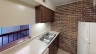 This two-floor residence offers a practical and comfortable layout with a series of well-defined living spaces. A defining architectural feature throughout the home is the exposed interior brick, which appears as a bold accent wall in multiple rooms, adding warmth and character to the overall aesthetic.

The living areas are finished with soft wall-to-wall carpeting and feature large sliding glass doors dressed with vertical blinds, allowing generous amounts of natural light to fill the space while providing views to the outdoors. Additional rooms include carpeted bedrooms with windows overlooking the surrounding neighborhood, each accented by the same signature brick wall detail.

The kitchen is equipped with dark wood cabinetry, a double-basin stainless steel sink set into light countertops, a white electric coil-burner range, a range hood, and a refrigerator. The brick wall extends into the kitchen, lending a cohesive rustic touch to the functional space.

The home includes two bathrooms: a full bathroom with a tub-shower combination, a vanity with a sink, and tile flooring, and a half bathroom with tile flooring, a vanity, and a toilet. Textured ceilings are present throughout the home, contributing to its classic residential character.