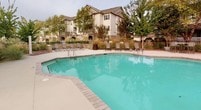 This single-story amenity space is designed to support an active lifestyle, combining a comprehensive fitness center with an inviting outdoor pool area. The fitness center is thoughtfully laid out across a multi-sided room, featuring coffered ceilings with warm wood trim detailing that lend an upscale, polished character to the space. Large sliding glass windows line multiple walls, flooding the interior with natural light and offering direct views of the outdoor pool and surrounding landscaped grounds. The facility is well-stocked with cardio equipment, including treadmills and elliptical machines positioned to take advantage of the pool views, alongside a robust selection of strength-training machines and free weights. Full-length mirrors amplify the sense of space, while rubberized flooring provides practical, durable underfoot support. A dedicated functional training area includes stability balls, medicine balls, resistance equipment, and a stationary bike, catering to a variety of workout styles. Outside, the pool area features a generously sized, irregularly shaped pool surrounded by a broad concrete deck. Poolside seating is available, and the area is enclosed by a wrought-iron fence. Mature trees and ornamental grasses add natural texture to the landscaping throughout the outdoor space.