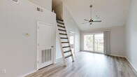 This 2-bedroom, 3-bathroom residence offers a thoughtfully designed layout across a single floor, highlighted by a striking architectural character throughout. The open-concept living room immediately captures attention with its soaring vaulted ceilings, a ceiling fan with a brass pendant fixture, and light wood-style flooring that extends seamlessly throughout the space. A unique loft-style staircase adds a distinctive architectural element, leading to an upper loft area visible from below. Large sliding glass doors open to a private balcony, flooding the living area with natural light and offering outdoor views of palm trees and the surrounding landscape. The kitchen features crisp white shaker cabinetry, light countertops, a stainless steel range with an overhead vent hood, and matching hardware, creating a clean and functional cooking space. The primary bedroom is generously proportioned, featuring a large window with plantation blinds that invite natural light. A dedicated walk-in closet provides ample storage with wire shelving and hanging rods. The additional bedroom is comfortably sized with light-filled windows. The bathrooms are finished with white vanities, light countertops, wood-style flooring, and tub-shower combinations. A hallway and closet complete the well-organized floor plan of this bright and airy residence.
