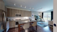 This single-floor residence showcases a thoughtfully designed open-concept layout that seamlessly connects the kitchen, dining, and living areas. The kitchen is a standout feature, outfitted with full-height wood-grain cabinetry, a suite of stainless steel appliances including a gas range, built-in microwave, refrigerator, and dishwasher, all complemented by sleek dark quartz countertops. A central island with bar seating provides both additional prep space and a casual dining option. Track lighting runs across the ceiling, offering bright, directed illumination throughout the cooking and living spaces. Rich hardwood flooring flows through the main living areas, adding warmth and continuity. The living room offers a comfortable, understated ambiance with soft neutral tones, and windows invite natural light into the space. The bedroom is a serene retreat featuring plush carpeting, multiple windows with views to the outdoors, and ample room for relaxation. The bathroom is finished with large-format tile flooring, a floating wood-toned vanity with a white countertop, a framed mirror, and modern lighting. Clean white walls and crisp millwork details throughout contribute to the residence's overall contemporary and welcoming character.