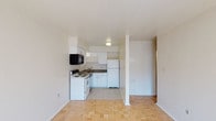 This single-floor unit presents a clean, functional layout with a seamless transition between the living area and kitchen. The kitchen is equipped with white cabinetry, a dark-toned countertop, a white electric range, a built-in black microwave, and a white refrigerator. A stainless sink is set beneath the upper cabinets, and light tile flooring defines the kitchen zone. The adjacent living area features warm parquet wood flooring in a classic basket-weave pattern, adding a touch of traditional character to the otherwise minimalist space. White walls and a white ceiling throughout the unit create a bright, airy atmosphere, enhanced by flush-mounted ceiling lighting. A structural column subtly divides the open-plan space without interrupting the overall flow. A hallway visible to the right suggests access to additional rooms or a bathroom, contributing to the efficient use of the floor plan. The overall ambiance is straightforward and practical, with a neutral palette that provides a versatile canvas for personalization.