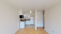 This single-floor unit presents a clean, functional layout with a seamless transition between the living area and kitchen. The kitchen is equipped with white cabinetry, a dark-toned countertop, a white electric range, a built-in black microwave, and a white refrigerator. A stainless sink is set beneath the upper cabinets, and light tile flooring defines the kitchen zone. The adjacent living area features warm parquet wood flooring in a classic basket-weave pattern, adding a touch of traditional character to the otherwise minimalist space. White walls and a white ceiling throughout the unit create a bright, airy atmosphere, enhanced by flush-mounted ceiling lighting. A structural column subtly divides the open-plan space without interrupting the overall flow. A hallway visible to the right suggests access to additional rooms or a bathroom, contributing to the efficient use of the floor plan. The overall ambiance is straightforward and practical, with a neutral palette that provides a versatile canvas for personalization.