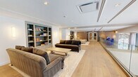 Located at 1860 Reston Row Plaza in Reston, Virginia, this thoughtfully designed single-floor space offers a refined and contemporary living environment. The layout centers around a generously sized living room that combines comfort and sophistication in equal measure.

Wide-plank hardwood flooring in a warm natural tone runs continuously throughout the space, anchoring the interior with an inviting warmth. A standout architectural feature is the sleek glass railing system, which adds a modern, open feel while maintaining visual connectivity across the space. The crisp white ceilings are adorned with recessed lighting and elegant crown molding details, contributing to a polished, upscale ambiance.

The living area is thoughtfully appointed with plush seating arrangements, including lounge chairs with ottomans set atop a classic area rug. Built-in bookshelves in a muted blue-gray finish provide functional storage and a curated aesthetic touch. Brass-finished floor lamps introduce a warm, ambient glow that complements the natural light filtering in through large windows with sheer drapery, offering views of the surrounding outdoor greenery.

The space also features a paneled accent wall in rich warm tones, adding depth and texture. An additional seating area is visible beyond the glass railing, further enhancing the spatial flow. Overall, the design strikes a balance between modern elegance and functional comfort.