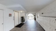 This single-floor residential building lobby offers a clean, functional, and well-maintained common area designed to serve its residents efficiently. The space is defined by its light-toned textured walls, which lend a warm, neutral character throughout. A standout architectural feature is the elegant arched entryway at the far end, framing a view to the outdoors and allowing natural light to filter into the corridor, creating an open and airy atmosphere.

The flooring consists of durable tile in a warm, earthy tone, providing a practical surface suited for high-traffic use. Recessed ceiling beams add a subtle structural detail overhead, complemented by flush-mounted ceiling lighting that ensures the corridor remains bright and welcoming.

On one side of the lobby, a bank of individual resident mailboxes lines the wall, offering convenient mail access in a compact, organized arrangement. Adjacent to the entrance is an elevator with wood-framed doors, providing vertical access throughout the building. Potted tropical plants are thoughtfully placed along the corridor, adding a touch of greenery and warmth to the otherwise utilitarian space. A doormat near the elevator reinforces the tidy, well-kept nature of the lobby. Overall, the space balances practicality with a hospitable ambiance.