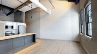 Unit 316 is a thoughtfully designed single-floor space that seamlessly blends industrial character with modern aesthetics. The open-concept layout creates a sense of spaciousness, anchored by soaring ceilings that showcase exposed ductwork, raw wooden beams, and bold white cylindrical pipes — all hallmarks of industrial-chic design. Light wood-tone luxury vinyl flooring runs throughout, lending warmth and continuity to the space. The kitchen is a standout feature, boasting dark wood cabinetry with a matte finish, a light-toned countertop, and a sleek gold-finish faucet at the sink. Pendant lights with matte black fixtures hang overhead, providing focused task lighting with a stylish edge. The open living area is bathed in natural light, with neutral gray walls offering a versatile backdrop that complements a variety of interior design styles. The combination of raw overhead elements with clean, polished surfaces below creates a compelling contrast that gives the space a dynamic and contemporary feel. This well-appointed unit offers a modern urban living experience with distinctive architectural details that set it apart.