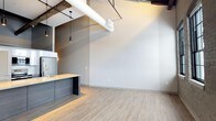 Unit 316 is a thoughtfully designed single-floor space that seamlessly blends industrial character with modern aesthetics. The open-concept layout creates a sense of spaciousness, anchored by soaring ceilings that showcase exposed ductwork, raw wooden beams, and bold white cylindrical pipes — all hallmarks of industrial-chic design. Light wood-tone luxury vinyl flooring runs throughout, lending warmth and continuity to the space. The kitchen is a standout feature, boasting dark wood cabinetry with a matte finish, a light-toned countertop, and a sleek gold-finish faucet at the sink. Pendant lights with matte black fixtures hang overhead, providing focused task lighting with a stylish edge. The open living area is bathed in natural light, with neutral gray walls offering a versatile backdrop that complements a variety of interior design styles. The combination of raw overhead elements with clean, polished surfaces below creates a compelling contrast that gives the space a dynamic and contemporary feel. This well-appointed unit offers a modern urban living experience with distinctive architectural details that set it apart.