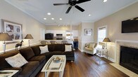 This single-floor home offers a thoughtfully designed open-concept layout that seamlessly connects the living, dining, and kitchen areas into one cohesive, comfortable space. Rich, dark hardwood floors flow throughout, anchoring the warm, neutral color palette of soft beige walls and crisp white trim. The living area is anchored by a large, plush dark sectional sofa paired with a sleek glass coffee table, creating an inviting gathering space. A ceiling fan with recessed lighting overhead ensures the area is both well-lit and comfortable year-round. A cozy accent chair and side table add an additional seating nook near the window, which allows natural light to filter in through slatted blinds. The kitchen is outfitted with classic white cabinetry, black appliances, and a breakfast bar with seating, making it both functional and stylish. Track lighting illuminates the kitchen workspace, while a window above the sink brings in additional natural light. Crown molding and chair rail detailing add a refined architectural touch throughout the space. The overall ambiance is warm, approachable, and well-appointed, balancing casual comfort with tasteful design elements that make the space feel both livable and polished.