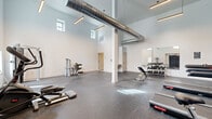 Located at 6822 Ridge Ave in the Roxborough neighborhood of Philadelphia, PA, this fitness center offers a spacious and thoughtfully designed workout environment across a single floor. The space is defined by its dramatically high ceilings, which lend an open, airy atmosphere throughout the main exercise area. Exposed industrial-style ductwork runs overhead, complementing the clean white walls and modern pendant lighting to create a contemporary yet functional aesthetic. High-set windows allow natural light to filter in, brightening the interior without compromising usable wall space. The main workout area features durable speckled rubber flooring, providing both safety and comfort underfoot. The space is well-appointed with a variety of equipment, including treadmills, stationary bikes, elliptical machines, adjustable benches, a dumbbell rack, cable machines, and step platforms, accommodating both cardio and strength training needs. Structural columns are incorporated seamlessly into the layout, and a large wall-mounted mirror on one side enhances the sense of depth and supports form-checking during workouts. A water fountain is conveniently mounted on the wall. The facility also includes a private restroom featuring a wood-toned vanity, full mirror, and tile flooring, ensuring a complete and convenient experience for users.