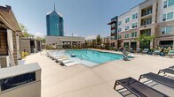 Located at 3007 Petty Rd in Durham, NC, this outdoor amenity space offers a well-appointed, resort-inspired environment designed for relaxation and community gathering. At its centerpiece is a generously sized swimming pool surrounded by a wide concrete deck lined with lounge chairs, providing ample space for sunbathing under the open North Carolina sky. A striking glass high-rise serves as a dramatic backdrop, lending an urban sophistication to the setting.

Adjacent to the pool, a manicured green lawn provides a versatile open-air retreat, accented by a pergola-style swing seating area ideal for casual lounging. Raised stone planters with native grasses, flowering shrubs, and ornamental plantings define pathways and add vibrant color throughout the grounds. A fire pit seating area offers an additional gathering spot, while private cabana structures with horizontal slatted siding and curtained interiors provide shaded, semi-enclosed retreats.

The surrounding multi-story building features a contemporary brick and panel facade with large, steel-framed grid windows that reflect the outdoor landscape and flood interior-facing spaces with natural light. Thoughtfully designed landscaping with river rock accents and layered plantings frames the building's exterior, creating a seamless transition between architecture and nature. The overall ambiance is modern, vibrant, and community-focused.