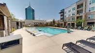 Located at 3007 Petty Rd in Durham, NC, this outdoor amenity space offers a well-appointed, resort-inspired environment designed for relaxation and community gathering. At its centerpiece is a generously sized swimming pool surrounded by a wide concrete deck lined with lounge chairs, providing ample space for sunbathing under the open North Carolina sky. A striking glass high-rise serves as a dramatic backdrop, lending an urban sophistication to the setting.

Adjacent to the pool, a manicured green lawn provides a versatile open-air retreat, accented by a pergola-style swing seating area ideal for casual lounging. Raised stone planters with native grasses, flowering shrubs, and ornamental plantings define pathways and add vibrant color throughout the grounds. A fire pit seating area offers an additional gathering spot, while private cabana structures with horizontal slatted siding and curtained interiors provide shaded, semi-enclosed retreats.

The surrounding multi-story building features a contemporary brick and panel facade with large, steel-framed grid windows that reflect the outdoor landscape and flood interior-facing spaces with natural light. Thoughtfully designed landscaping with river rock accents and layered plantings frames the building's exterior, creating a seamless transition between architecture and nature. The overall ambiance is modern, vibrant, and community-focused.