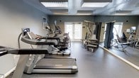 This fitness center offers a comprehensive workout environment designed to meet a wide range of exercise needs. The space is finished with a durable, speckled rubber flooring that provides both safety and comfort underfoot, ideal for high-traffic gym use. The walls are painted in a cool, neutral gray tone, lending the room a focused and energizing atmosphere.

Overhead, large recessed panel lights illuminate the entire space evenly, ensuring excellent visibility throughout all workout areas. Natural light also filters in through a set of double French doors fitted with shuttered blinds, adding a welcoming brightness to the room.

The cardio section is prominently featured with a row of Matrix-brand treadmills and elliptical trainers, offering users professional-grade equipment for endurance and cardiovascular training. Toward the back of the room, a multi-station strength training machine provides versatile options for resistance and muscle-building exercises, complementing the cardio offerings with a full-body workout capability.

The layout is thoughtfully arranged to allow for comfortable movement between machines, and the overall design prioritizes functionality without sacrificing a clean, polished aesthetic. This fitness center is well-suited for regular, varied exercise routines in a professional and well-maintained setting.