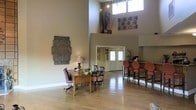 This single-floor space makes a strong first impression with its dramatic double-height ceilings that lend an open, airy atmosphere to the interior. Natural light streams in through tall clerestory windows positioned near the top of the walls, complemented by track lighting mounted along the upper ceiling to illuminate the space effectively at any time of day.

The main area features warm-toned hardwood flooring throughout, contributing to an inviting and cohesive aesthetic. A wooden desk with cross-back chairs serves as a central reception or workspace area, accented by a table lamp and floral arrangement that add a welcoming touch. Adjacent to the desk, a bar-height counter with upholstered rust-orange stools provides an additional seating area, well-suited for casual interaction or light refreshments.

Decorative wall art is thoughtfully placed throughout, including a large textured wall piece and an ornamental panel, enhancing the visual character of the space. Tall decorative vases with branches are displayed on an elevated ledge, adding vertical interest that draws the eye upward. Glass double doors at the far end of the space provide natural light and easy access to the exterior. The overall ambiance is warm, functional, and professionally appointed.
