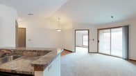 This single-floor unit offers a well-designed, open-concept layout that efficiently connects the main living areas. The kitchen is outfitted with warm medium-toned wood cabinetry, granite-style countertops, and a full suite of black appliances, including a refrigerator, range, microwave, and dishwasher. Hardwood-style flooring runs through the kitchen, which opens to the living and dining areas via a peninsula breakfast bar with pendant lighting above. The living area transitions to plush carpeting and features a sliding glass door leading to a private balcony, filling the space with natural light and outdoor views. Multiple bedrooms are carpeted and each includes a ceiling fan with lighting and a window for natural light. One room features a large picture window with views of the surrounding neighborhood. The bathroom includes a full tub and shower combination with clean white surround. Throughout the unit, warm wood-toned trim and doors provide consistent design continuity. The overall layout, as seen in the floorplan, is thoughtfully arranged to maximize usability across all areas of the home.