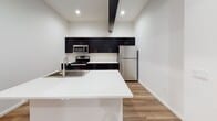 This modern single-floor unit showcases a thoughtfully designed open-concept layout that seamlessly connects the living area to the kitchen. The living space features warm wood-look flooring, crisp white walls, and exposed ductwork that lends an industrial-chic character, complemented by statement pendant lighting overhead.

The kitchen is well-appointed with dark, flat-front cabinetry, bright white countertops, a large kitchen island with a built-in sink, and a full suite of stainless steel appliances including a refrigerator, electric range, and microwave. Recessed lighting keeps the kitchen bright and functional.

The bedrooms offer plush carpeting and generous natural light through large windows fitted with blinds, providing outdoor views of the surrounding area. Each bedroom also features built-in closet shelving for organized storage.

The bathrooms are finished in a clean, contemporary style with wood-look tile flooring, dark vanity cabinetry, white countertops with dual sinks, large mirrors, and chrome fixtures. A walk-in shower with built-in shelving completes the primary bath.

Overall, the unit presents a cohesive modern aesthetic with a neutral palette, balancing industrial design elements with clean, refined finishes throughout.