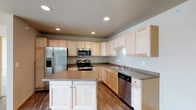 This single-story space is thoughtfully designed to maximize functionality and flow across its open layout. The floor plan reveals distinct living, dining, and kitchen zones that connect seamlessly, creating a cohesive and practical living environment.

The kitchen serves as a central highlight of the space, outfitted with light maple cabinetry that extends to the ceiling, providing generous storage throughout. Granite-style countertops run along the perimeter and continue onto a freestanding central island, which offers additional prep space and casual seating potential. Stainless steel appliances, including a French door refrigerator with an ice and water dispenser, a range with oven, an over-the-range microwave, and a dishwasher, equip the kitchen for everyday use. Recessed lighting illuminates the space evenly, while hardwood-style flooring ties the kitchen to the adjacent living areas.

The living areas are carpeted in sections, offering a comfortable contrast to the hard-surface flooring in the kitchen and dining zones. Natural light filters in through windows positioned along the exterior walls, contributing to a bright and welcoming atmosphere throughout the single-floor layout.