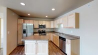 This single-story space is thoughtfully designed to maximize functionality and flow across its open layout. The floor plan reveals distinct living, dining, and kitchen zones that connect seamlessly, creating a cohesive and practical living environment.

The kitchen serves as a central highlight of the space, outfitted with light maple cabinetry that extends to the ceiling, providing generous storage throughout. Granite-style countertops run along the perimeter and continue onto a freestanding central island, which offers additional prep space and casual seating potential. Stainless steel appliances, including a French door refrigerator with an ice and water dispenser, a range with oven, an over-the-range microwave, and a dishwasher, equip the kitchen for everyday use. Recessed lighting illuminates the space evenly, while hardwood-style flooring ties the kitchen to the adjacent living areas.

The living areas are carpeted in sections, offering a comfortable contrast to the hard-surface flooring in the kitchen and dining zones. Natural light filters in through windows positioned along the exterior walls, contributing to a bright and welcoming atmosphere throughout the single-floor layout.