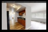 This single-floor, 2-bedroom, 1-bathroom unit offers a practical and efficiently arranged layout across a comfortable footprint. The living room is bright and open, featuring light wood-style laminate flooring, a wide window that invites natural light, a baseboard heating system, and a wall-mounted air conditioning unit for year-round climate control.

The kitchen is positioned adjacent to the living area through an arched opening, showcasing warm medium-toned wood cabinetry, dark stone-look countertops, and an integrated sink. The design creates a connected flow between the cooking and living spaces.

Both bedrooms are carpeted in neutral gray tones and are equipped with wall-mounted air conditioning units, baseboard heaters, and windows overlooking outdoor greenery. Each bedroom includes a closet with hanging rods for storage. The primary bedroom offers a comfortable, well-proportioned space with its own closet.

A hallway connects the main living spaces to the bedrooms and shared bathroom. The bathroom features a full soaking tub with a shower, complemented by bold, earth-toned large-format tile surround in warm brown and gray hues, adding a distinctive visual element to the space.

Overall, the unit presents a clean, functional design with consistent neutral tones throughout, practical storage solutions, and adequate natural light in all main living areas.