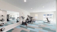Located at 446 Waterman Ave in East Providence, Rhode Island, this fitness center offers a well-appointed and functional space for residents to maintain an active lifestyle. The room is finished with a distinctive multi-toned geometric carpet in shades of teal, blue, and beige, adding a modern aesthetic to the clean, neutral-toned interior.

The cardio zone is well-equipped with two treadmills, an elliptical trainer, and a stationary bike, providing a variety of aerobic workout options. A multi-function cable resistance machine serves as the centerpiece for strength training, while a dedicated free weight rack stocked with dumbbells rounds out the equipment selection. Large wall-mounted mirrors expand the sense of space and allow users to monitor their form during workouts.

Natural light enters through multiple windows, creating a bright and energizing atmosphere. Recessed ceiling lighting ensures the space remains well-lit throughout the day. The room is accessed through a teal-framed glass door and is equipped with a fire alarm and emergency exit signage, reflecting attention to safety. The overall layout is open and spacious, allowing comfortable movement between equipment stations and accommodating multiple users simultaneously.