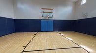 Located in Westworth Village, Texas, this single-floor indoor basketball facility offers a well-appointed court designed for athletic performance and safety. The court features a classic light maple hardwood-style tile floor with regulation court markings, including key areas, three-point arcs, and a center circle, providing a professional playing surface suitable for organized games and recreational play alike.

The perimeter walls are lined with thick navy blue padding that extends to approximately mid-wall height, offering essential protection for players. Above the padding, the walls transition to a clean white finish, contributing to a bright and open atmosphere. Each end of the court is equipped with a wall-mounted basketball hoop featuring a clear acrylic backboard, positioned at regulation height.

The facility boasts impressively high ceilings that accommodate full-court play without restriction. A row of clerestory windows positioned near the ceiling allows natural light to filter in from multiple sides of the court, supplementing the overhead ventilation and lighting systems. Access points are located along the lower padded walls, with clearly marked exit doors ensuring safe egress. The overall design prioritizes functionality, safety, and player comfort, making this a versatile indoor sports venue.