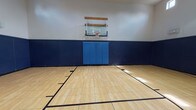 Located in Westworth Village, Texas, this single-floor indoor basketball facility offers a well-appointed court designed for athletic performance and safety. The court features a classic light maple hardwood-style tile floor with regulation court markings, including key areas, three-point arcs, and a center circle, providing a professional playing surface suitable for organized games and recreational play alike.

The perimeter walls are lined with thick navy blue padding that extends to approximately mid-wall height, offering essential protection for players. Above the padding, the walls transition to a clean white finish, contributing to a bright and open atmosphere. Each end of the court is equipped with a wall-mounted basketball hoop featuring a clear acrylic backboard, positioned at regulation height.

The facility boasts impressively high ceilings that accommodate full-court play without restriction. A row of clerestory windows positioned near the ceiling allows natural light to filter in from multiple sides of the court, supplementing the overhead ventilation and lighting systems. Access points are located along the lower padded walls, with clearly marked exit doors ensuring safe egress. The overall design prioritizes functionality, safety, and player comfort, making this a versatile indoor sports venue.
