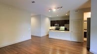 This single-floor space offers a clean, modern aesthetic defined by an open-concept layout that seamlessly connects the living area to the kitchen. Rich hardwood flooring runs throughout, adding warmth and continuity to the open floor plan. The living area is generously proportioned, with smooth white walls, recessed lighting, and a track lighting fixture that provides ample illumination across the space.

The kitchen is a standout feature, equipped with dark espresso cabinetry that contrasts elegantly against a light-toned countertop. A peninsula with a breakfast bar separates the kitchen from the living area, creating a natural flow while maintaining an open feel. Stainless steel appliances — including a refrigerator, microwave, and range — lend a polished, contemporary look. A green glass tile backsplash adds a vibrant accent and visual interest against the dark cabinetry.

Additional details include a louvered door offering practical storage access, recessed ceiling vents, and thoughtful lighting placement throughout. The overall ambiance is bright, airy, and move-in ready, making it a highly functional and visually cohesive space well-suited for modern living.