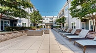 Located at 2142 Commonwealth Ave in Charlotte's vibrant 28205 corridor, this expansive outdoor courtyard amenity space is nestled within a multi-story residential community designed with a contemporary aesthetic. The building's gray lap-siding exterior, large windows, and private balconies frame the courtyard on multiple sides, creating an enclosed, resort-like atmosphere open to the sky.

The generously sized paved terrace is finished with large-format stone tiles throughout, providing a clean, cohesive look across the entire outdoor area. Along one side, a row of cushioned chaise lounge chairs invites residents to relax and enjoy natural sunlight, while mature trees interspersed throughout the courtyard offer natural shade and greenery. Multiple seating vignettes featuring wicker and upholstered outdoor sofas are arranged around central gathering areas, ideal for socializing or quiet relaxation.

A prominent pergola structure anchors the center of the courtyard, providing a shaded focal point and defined outdoor entertaining zone. A secondary gazebo with a pitched roof and integrated lighting adds further architectural interest and functionality. Raised stone planter beds line portions of the courtyard perimeter, introducing layered landscaping that softens the hardscape. String lights are strung overhead, enhancing the ambiance during evening hours. Gated access points connect the courtyard directly to individual building entrances, ensuring both convenience and security.