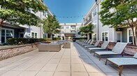 Located at 2142 Commonwealth Ave in Charlotte's vibrant 28205 corridor, this expansive outdoor courtyard amenity space is nestled within a multi-story residential community designed with a contemporary aesthetic. The building's gray lap-siding exterior, large windows, and private balconies frame the courtyard on multiple sides, creating an enclosed, resort-like atmosphere open to the sky.

The generously sized paved terrace is finished with large-format stone tiles throughout, providing a clean, cohesive look across the entire outdoor area. Along one side, a row of cushioned chaise lounge chairs invites residents to relax and enjoy natural sunlight, while mature trees interspersed throughout the courtyard offer natural shade and greenery. Multiple seating vignettes featuring wicker and upholstered outdoor sofas are arranged around central gathering areas, ideal for socializing or quiet relaxation.

A prominent pergola structure anchors the center of the courtyard, providing a shaded focal point and defined outdoor entertaining zone. A secondary gazebo with a pitched roof and integrated lighting adds further architectural interest and functionality. Raised stone planter beds line portions of the courtyard perimeter, introducing layered landscaping that softens the hardscape. String lights are strung overhead, enhancing the ambiance during evening hours. Gated access points connect the courtyard directly to individual building entrances, ensuring both convenience and security.