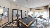 This well-appointed billiard room serves as a dynamic social space that seamlessly blends recreation with casual functionality. At its center sits a full-size pool table with a slate-blue felt surface and a sleek modern frame, positioned on a bold geometric-patterned carpet that anchors the room with visual energy. A wall-mounted cue rack keeps equipment neatly organized and within easy reach.

Natural light fills the space through large windows fitted with roller shades and sheer curtain panels, offering views of an outdoor pool area beyond. This connectivity to the exterior gives the room an open, airy quality despite its defined footprint.

Along one side, a dedicated seating and meeting area features two square tables surrounded by contemporary task chairs on casters, making it a practical spot for informal gatherings or collaborative use. A curated gallery wall of framed artwork adds a refined, residential touch to the neutral warm-gray walls, while recessed ceiling lighting provides consistent, even illumination throughout.

French doors with dark frames lead directly to an adjacent outdoor or common area, enhancing the room's accessibility and flow. A potted plant and additional art pieces contribute to the polished yet inviting atmosphere. The overall design balances active recreation with comfortable social interaction in a cohesive, modern aesthetic.