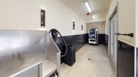 This dog wash facility is designed with both functionality and durability in mind. The interior features a consistent design language throughout, with dark vertically-laid tile wainscoting lining the lower half of all walls, contrasting against cream-colored upper walls. The flooring is polished concrete, chosen for its easy-clean practicality in a wet environment.

The main washing area is equipped with stainless steel bathing tubs, providing a hygienic and sturdy surface for pet washing. A dedicated hot water heater ensures a consistent warm water supply, while a built-in hose and dryer attachment offer convenience for a complete grooming session. Floor-level metal grating in the wash area facilitates efficient drainage.

The space is well-lit with overhead fluorescent fixtures and supplemented by modern wall-mounted sconces. Large windows and glass-paneled double doors allow natural light to flow in, offering views of an outdoor covered pergola structure. The facility also includes a vending machine, adding convenience for users. The overall layout is open and elongated, providing ample room to maneuver, and the finishes throughout are clearly selected for longevity and ease of maintenance.