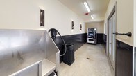 This dog wash facility is designed with both functionality and durability in mind. The interior features a consistent design language throughout, with dark vertically-laid tile wainscoting lining the lower half of all walls, contrasting against cream-colored upper walls. The flooring is polished concrete, chosen for its easy-clean practicality in a wet environment.

The main washing area is equipped with stainless steel bathing tubs, providing a hygienic and sturdy surface for pet washing. A dedicated hot water heater ensures a consistent warm water supply, while a built-in hose and dryer attachment offer convenience for a complete grooming session. Floor-level metal grating in the wash area facilitates efficient drainage.

The space is well-lit with overhead fluorescent fixtures and supplemented by modern wall-mounted sconces. Large windows and glass-paneled double doors allow natural light to flow in, offering views of an outdoor covered pergola structure. The facility also includes a vending machine, adding convenience for users. The overall layout is open and elongated, providing ample room to maneuver, and the finishes throughout are clearly selected for longevity and ease of maintenance.