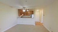 This single-floor space is designed with an open-concept layout that integrates the kitchen and living area into one cohesive environment. The kitchen is defined by warm honey-toned wood cabinetry that provides ample storage, complemented by granite-look countertops and a stainless steel refrigerator. A raised peninsula separates the kitchen from the living area, offering a practical surface for casual dining or meal prep while maintaining visual openness throughout the space.

The living area features plush carpeting, which contrasts with the light hardwood flooring found in the kitchen and hallway beyond. A ceiling fan with an integrated light fixture is centrally positioned, providing both air circulation and ambient lighting. The neutral, light-colored walls throughout contribute to a bright and airy atmosphere.

A hallway visible from the main living space leads to additional rooms, with a doorway at the end suggesting further private quarters. The layout flows naturally from the living and kitchen areas toward these spaces, making the overall floor plan feel functional and well-organized. The clean, understated design aesthetic gives the space a versatile and welcoming character.