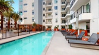 This outdoor amenity space is centered around a generously sized swimming pool set within a landscaped courtyard enclosed by a contemporary multi-story building. The pool deck is finished in light-toned concrete and lined with rows of sleek, dark-framed chaise lounges fitted with cushioned pads, providing ample space for sunbathing. Multiple seating arrangements, including modular outdoor sofas with navy blue cushions and low coffee tables, are positioned throughout the deck to create distinct relaxation zones. Bold terracotta-orange planters of varying sizes serve as a defining design element throughout the space, housing lush tropical plants including bird-of-paradise and banana leaf varieties, lending vivid color contrast against the building's crisp white and neutral facades. The courtyard opens to sky views framed by towering palm trees, reinforcing a warm, Southern California aesthetic. A black iron perimeter fence encloses the pool area for safety. Overhead, the surrounding building features private balconies with metal railings that overlook the courtyard. Additional raised planter beds along walkways introduce further greenery. The overall ambiance is modern, resort-inspired, and light-filled.