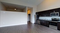This single-floor residence offers a well-designed open-concept layout that seamlessly blends living and kitchen areas into one cohesive space. Rich, dark hardwood flooring runs throughout, providing a warm and contemporary foundation that complements the clean, neutral-toned walls.

The kitchen is thoughtfully appointed with dark espresso cabinetry offering generous storage, granite countertops, and stainless steel appliances including a refrigerator and dishwasher. A double-basin sink sits beneath a classic subway tile backsplash, completing the modern aesthetic.

The adjoining living area is bright and open, featuring elevated ceilings that enhance the sense of spaciousness. A ceiling fan adds both function and style to this comfortable gathering space. The open floor plan creates a natural flow between the kitchen and living zones, making the space feel expansive and connected.

A doorway visible from the main living area leads to additional spaces, hinting at a walk-in closet or further rooms beyond. The clean architectural lines, consistent flooring, and cohesive color palette give this residence a polished, move-in-ready appeal. The overall ambiance is modern, clean, and functional — ideal for everyday living.
