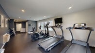 Located at 35 Sharon Rd in Waterbury, Connecticut, this fitness center offers a compact yet efficiently designed space for residents or members to maintain an active lifestyle. The single-floor layout includes a dedicated exercise room and two restrooms.

The exercise room is the heart of this facility, featuring a dark rubber flooring that provides both durability and comfort during workouts. The space is outfitted with multiple treadmills, an elliptical trainer, a stationary bike, and a weight bench with dumbbells, offering a well-rounded cardio and strength training experience. A wall-mounted flat-screen television keeps users entertained during workouts. Natural light streams in through several windows, creating a bright and energizing atmosphere. The walls are finished in a two-tone scheme of cool gray and white, complemented by recessed ceiling lighting for a clean, modern look. A wall-mounted mirror on one side enhances the sense of space. A climate control unit ensures year-round comfort.

The two restrooms are straightforward and functional, featuring wood-look vinyl flooring and textured white walls. One restroom is designed as a men's facility with a stall, urinal, and wall-mounted sink, while the other offers a similar practical layout, both equipped with soap dispensers and paper towel fixtures for convenience.