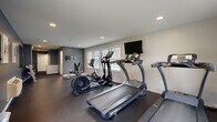 Located at 35 Sharon Rd in Waterbury, Connecticut, this fitness center offers a compact yet efficiently designed space for residents or members to maintain an active lifestyle. The single-floor layout includes a dedicated exercise room and two restrooms.

The exercise room is the heart of this facility, featuring a dark rubber flooring that provides both durability and comfort during workouts. The space is outfitted with multiple treadmills, an elliptical trainer, a stationary bike, and a weight bench with dumbbells, offering a well-rounded cardio and strength training experience. A wall-mounted flat-screen television keeps users entertained during workouts. Natural light streams in through several windows, creating a bright and energizing atmosphere. The walls are finished in a two-tone scheme of cool gray and white, complemented by recessed ceiling lighting for a clean, modern look. A wall-mounted mirror on one side enhances the sense of space. A climate control unit ensures year-round comfort.

The two restrooms are straightforward and functional, featuring wood-look vinyl flooring and textured white walls. One restroom is designed as a men's facility with a stall, urinal, and wall-mounted sink, while the other offers a similar practical layout, both equipped with soap dispensers and paper towel fixtures for convenience.