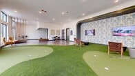 Located at 5720 Peachtree Pkwy in Peachtree Corners, GA, this single-floor recreational space offers a generously sized, thoughtfully designed golf and entertainment lounge. The centerpiece is a multi-hole indoor putting green with contoured turf surfaces, providing an engaging golf experience without stepping outside. Adjacent to the putting green is a dedicated golf simulator bay, enclosed with dark curtaining and equipped with a projection screen and computer setup running a virtual golf platform, allowing players to experience full-course play year-round.

The space transitions smoothly between the artificial turf areas and wood-look tile flooring, creating distinct zones for play and socializing. Unique suspended rope swings with wooden bench seats add a playful, distinctive design element, while high-top tables and modern chairs offer casual seating for spectators or groups. A decorative arched accent wall with bold patterned wallcovering frames a mounted display screen, adding visual character to the interior.

Large floor-to-ceiling windows line the perimeter walls, flooding the space with natural light and offering views of the surrounding landscape. Recessed ceiling lighting provides consistent illumination throughout. The overall ambiance is modern, welcoming, and purpose-built for golf enthusiasts and social gatherings alike.
